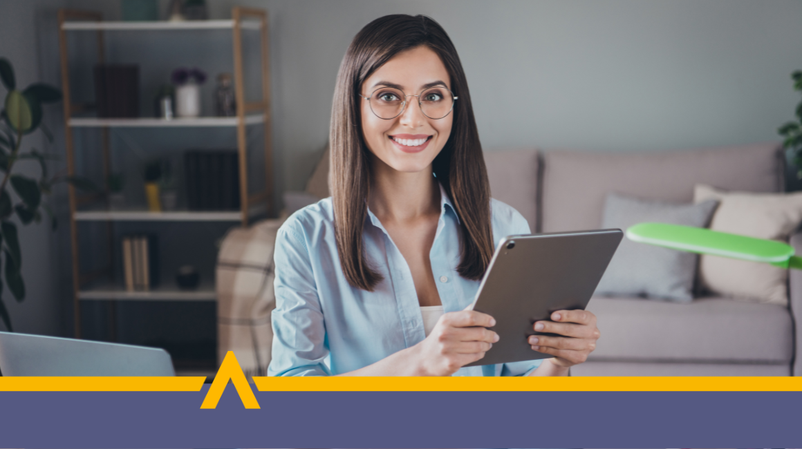 Młoda kobieta w okularach i błękitnej koszuli trzyma tablet, siedzi przy biurku z laptopem, w tle nowoczesne wnętrze z regałem i sofą